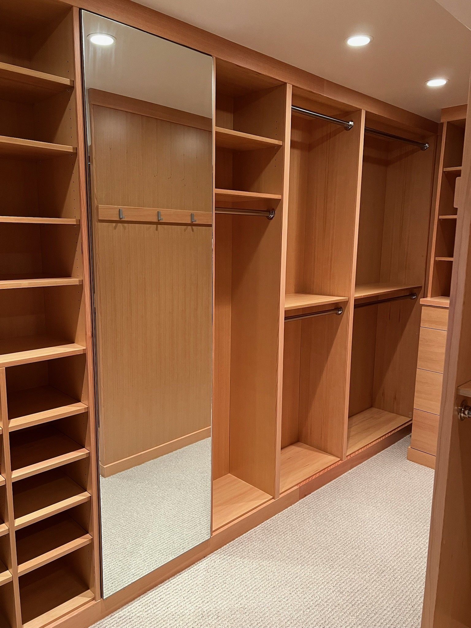 Wooden walk-in closet with shelves, hanging rods, and full-length mirror. Light-colored wood against a neutral carpet.