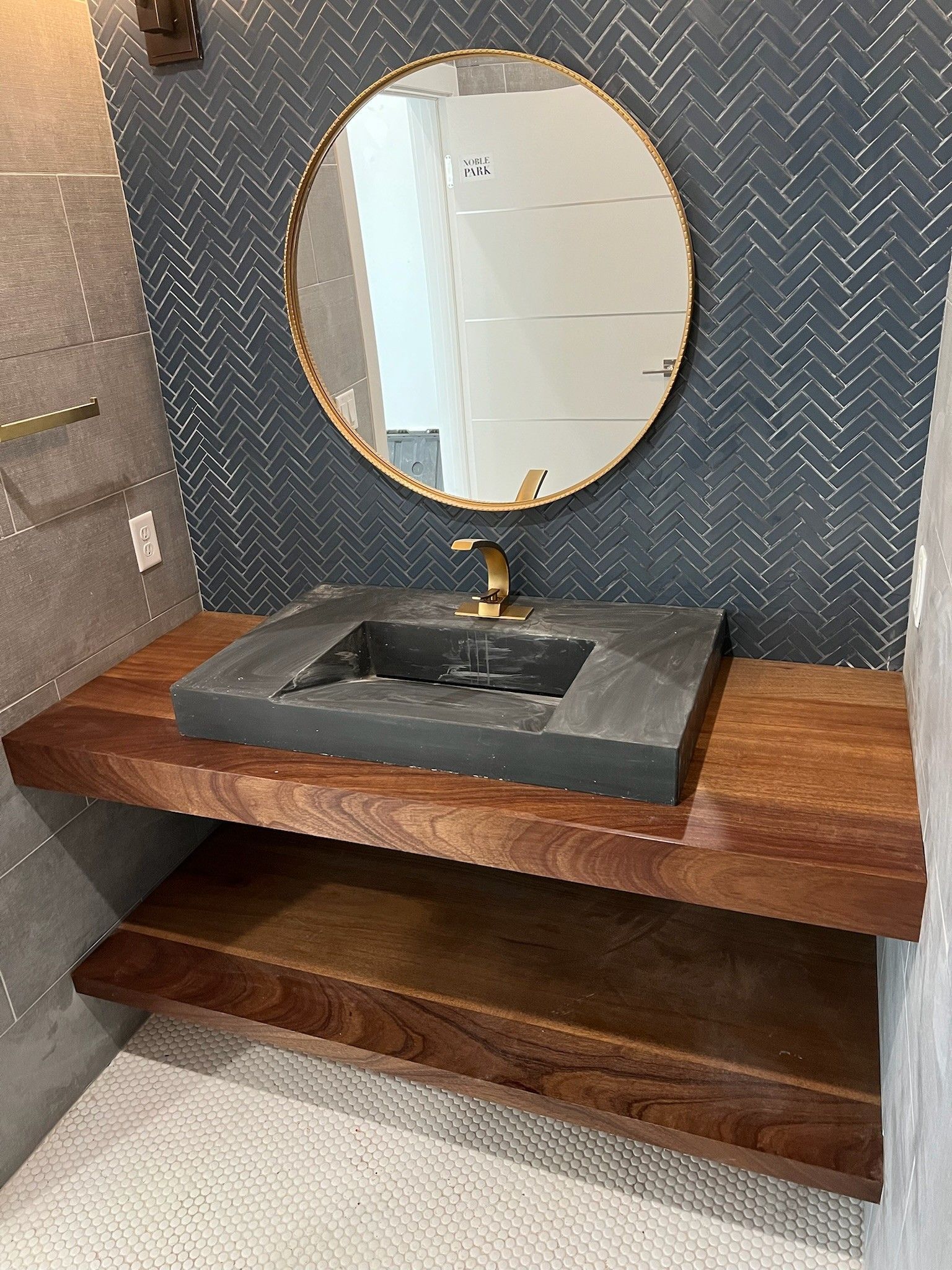 Bathroom with a floating wood shelf, black sink, gold faucet, and circular mirror. Dark blue tiled backsplash.