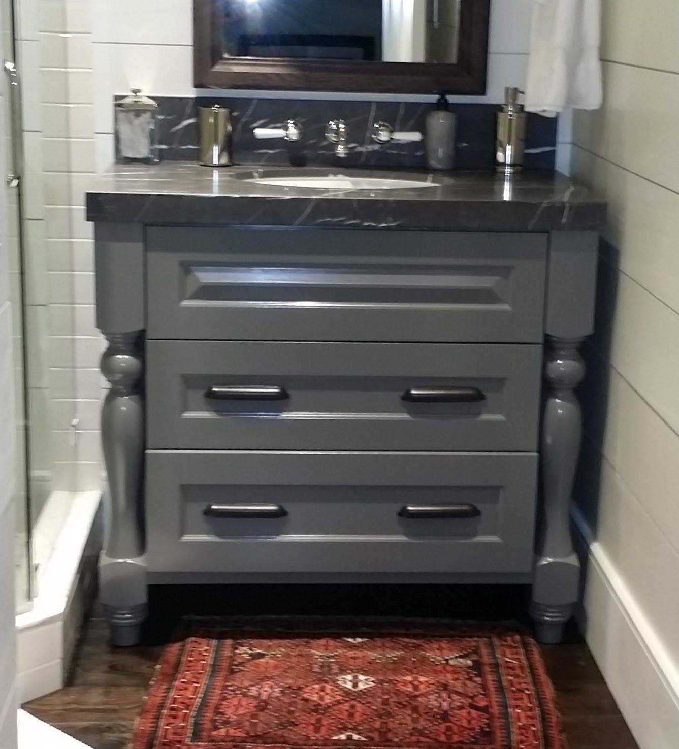 Gray vanity with black countertop, two drawers, and ornate legs.  Small rug, and dark-framed mirror above.