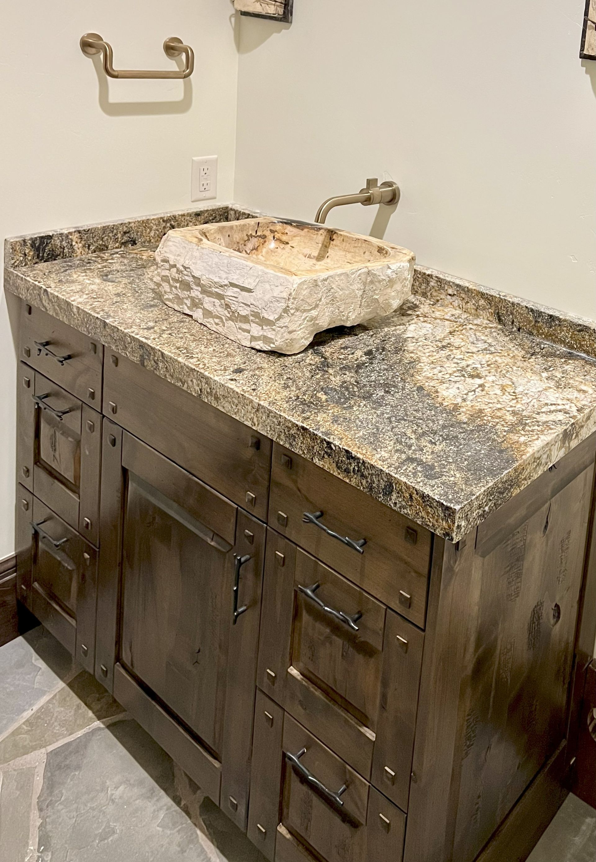 Rustic bathroom vanity with stone sink and granite countertop. Wooden cabinet, brass fixtures, and stone floor.