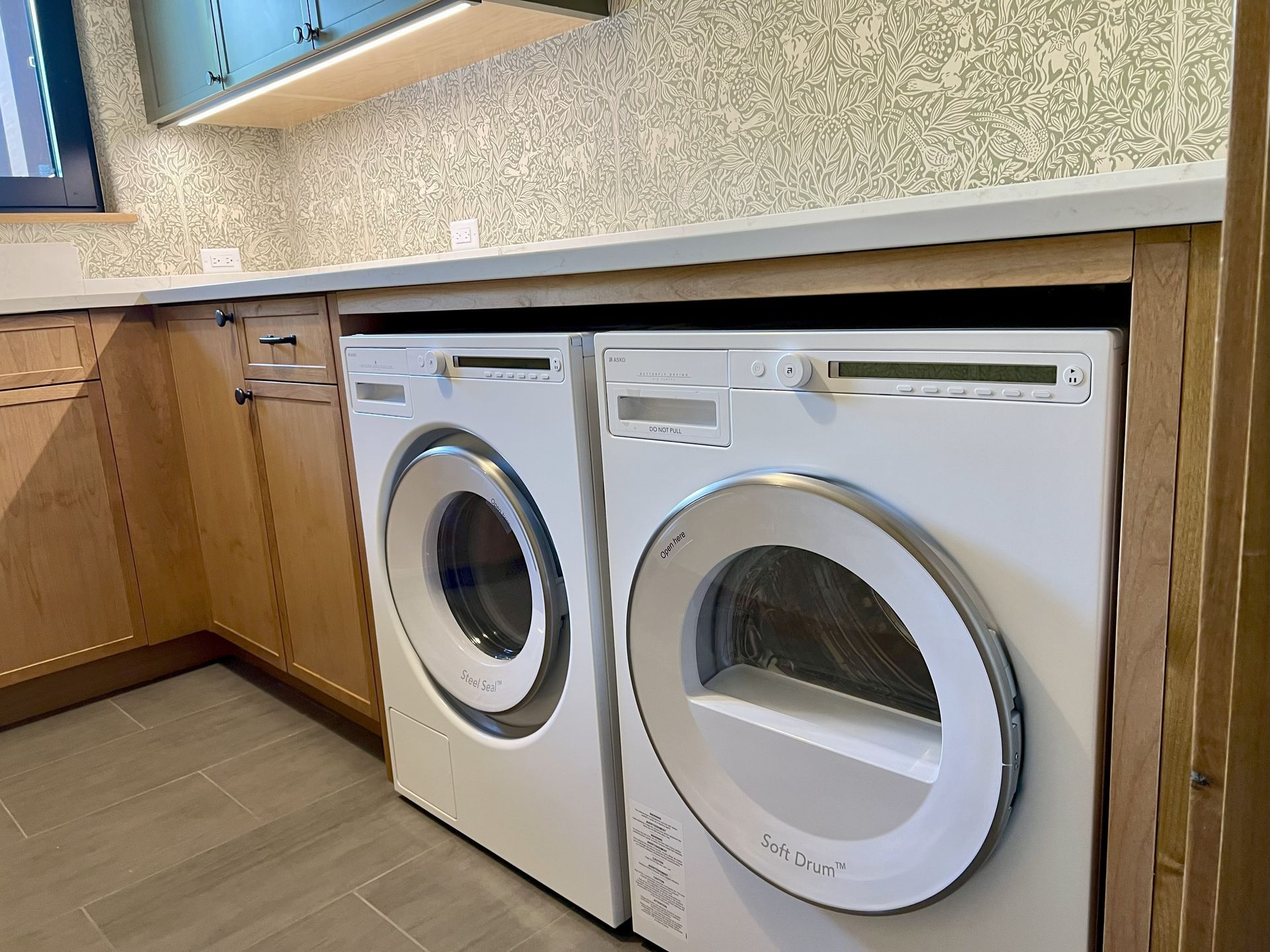 Washer and dryer set, white with silver trim, in a laundry room with wooden cabinetry and light countertops.