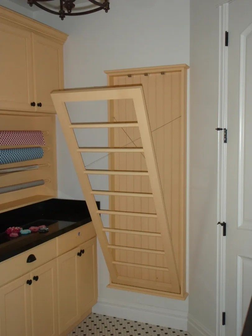Fold-down wooden drying rack on a wall in a laundry room, partially opened, beige cabinets, black countertop, and a white door.