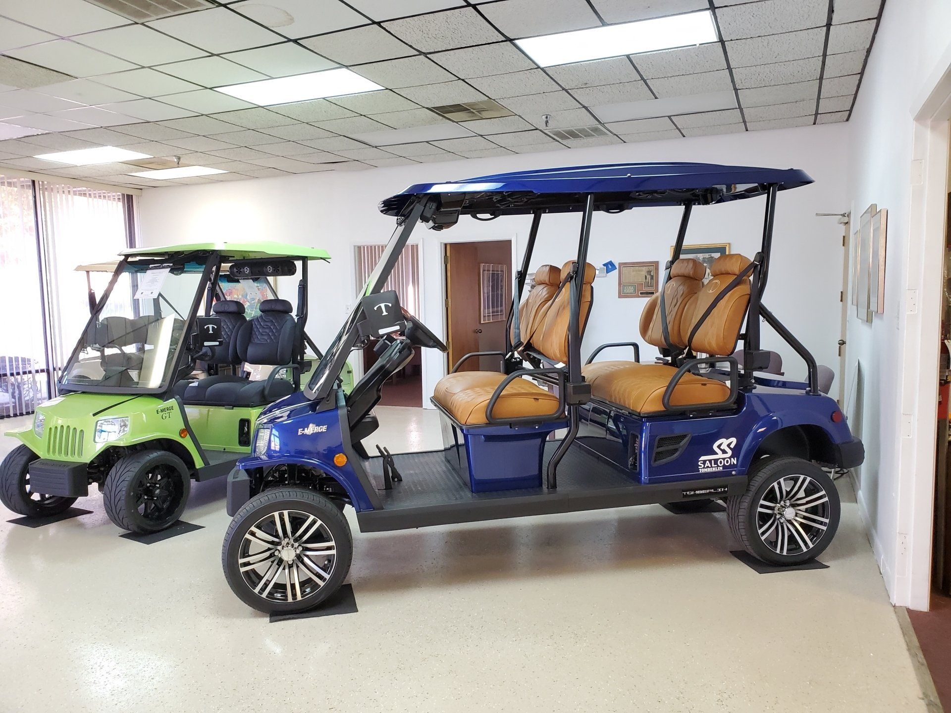 Two golf carts, one green and one blue, on display inside a showroom. The blue cart has tan seats.