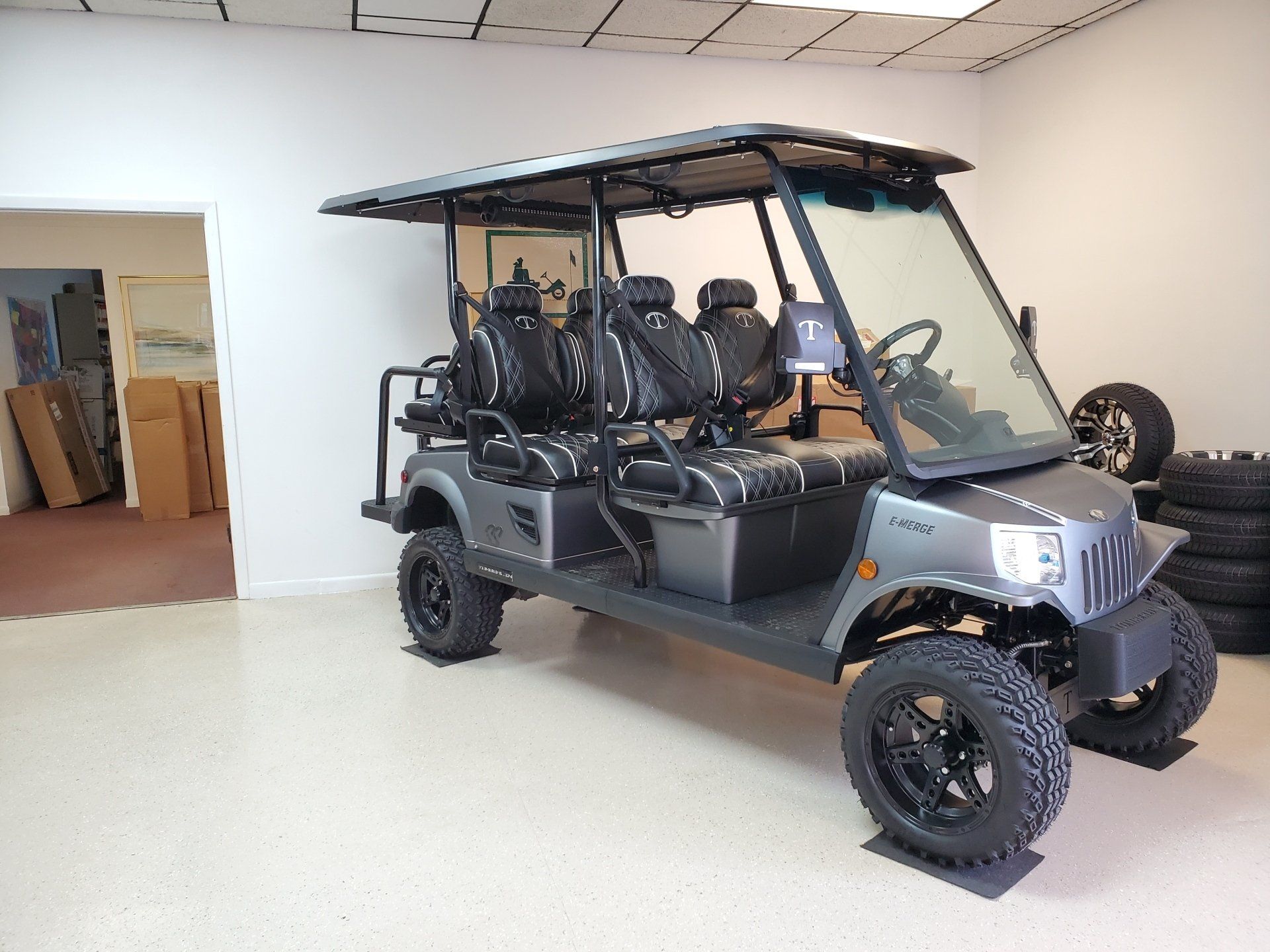 A gray, six-passenger golf cart parked indoors. It has black seats, black wheels, and a canopy.