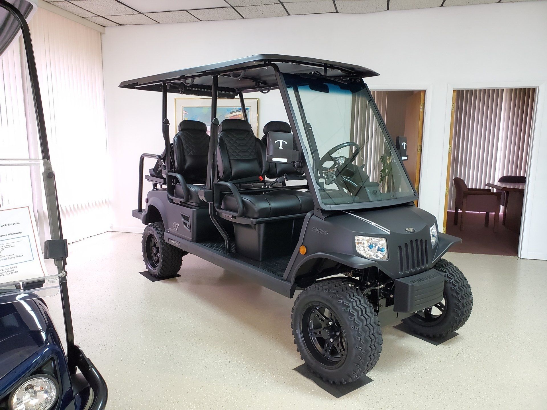 Black golf cart with off-road tires inside a showroom. It has a roof, windshield, and seating for multiple passengers.