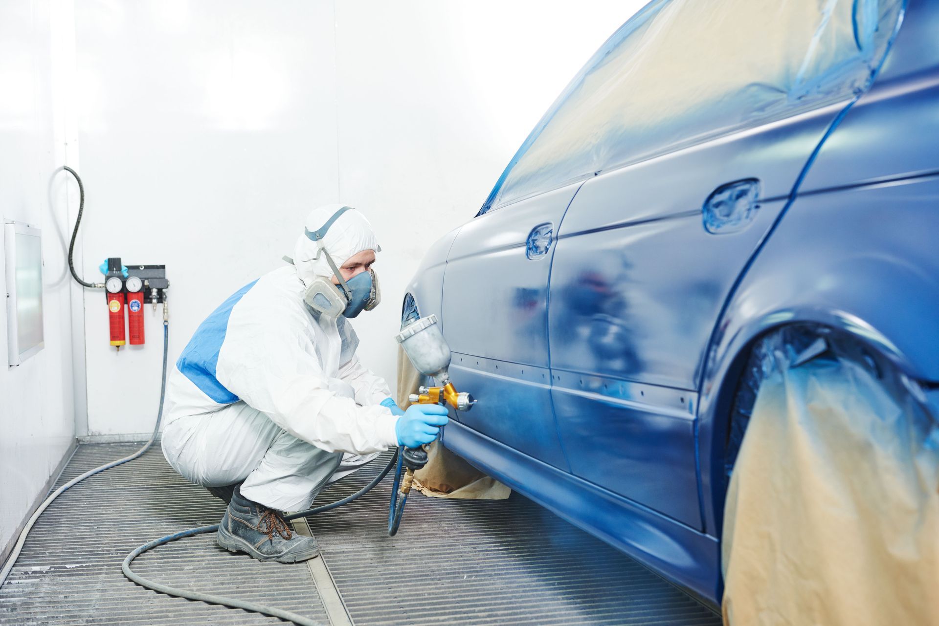 Person in protective suit spray-painting a blue car in a paint booth.