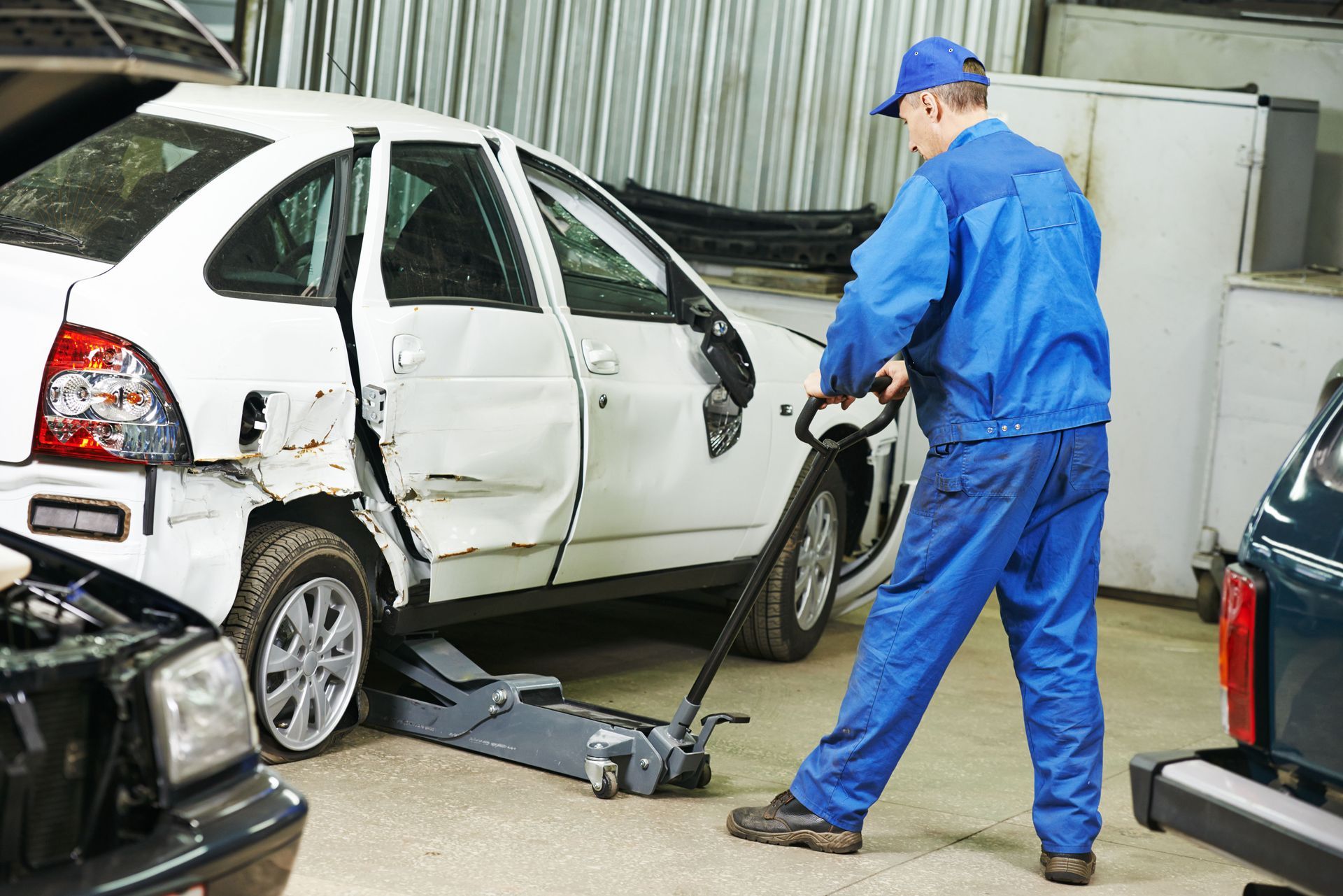 Mechanic uses a jack to lift a damaged white car in a garage.