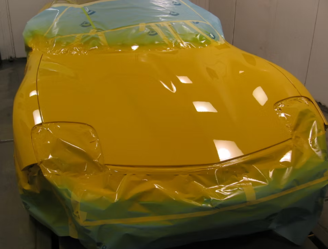 Yellow car hood covered in protective plastic in a paint booth.