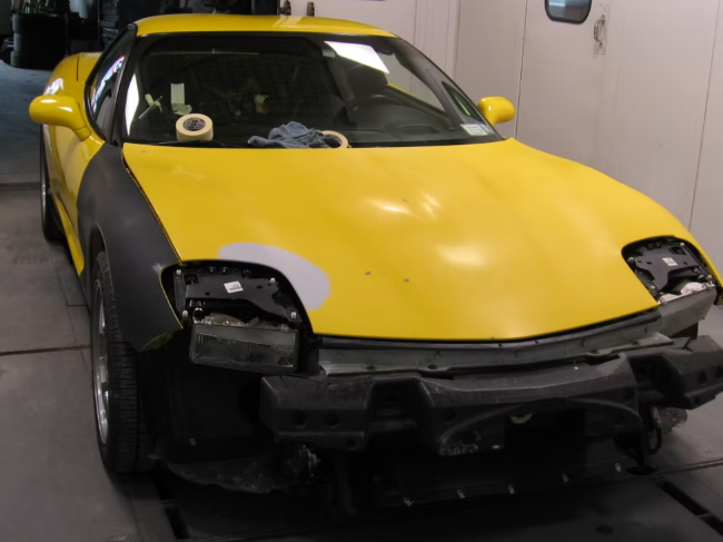 Yellow Corvette undergoing repair in a shop; missing headlights and front bumper.