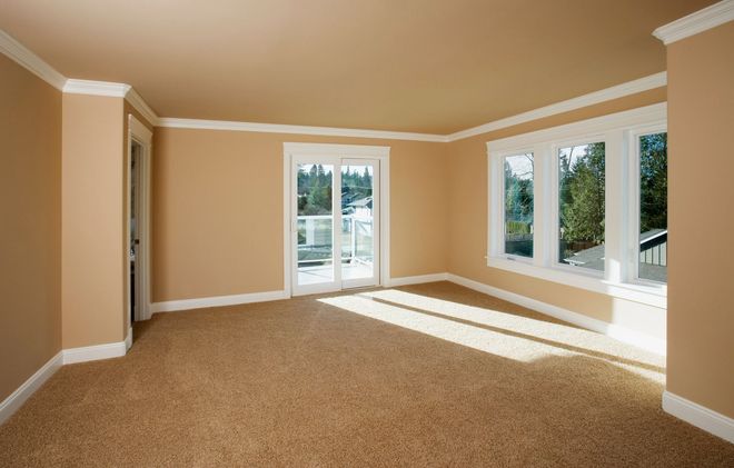 Empty room with tan carpet, walls, and white trim. Sliding door and windows.