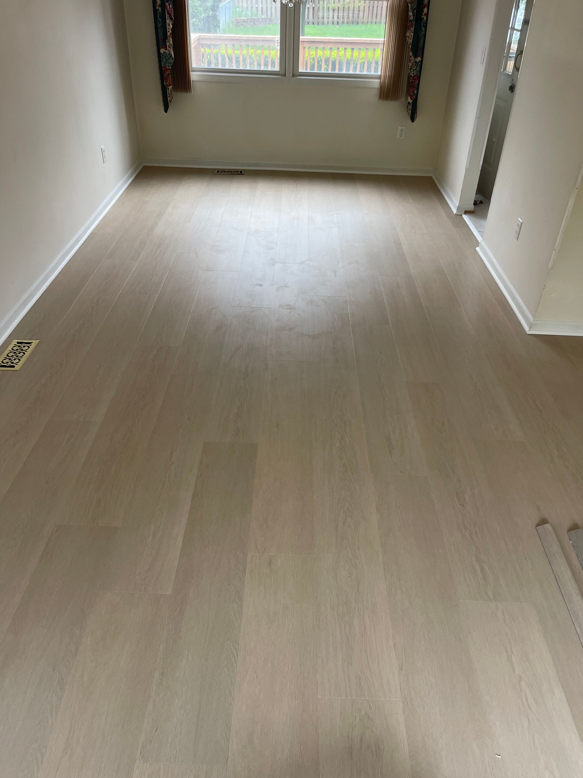 Light wood flooring in a room with a window. The walls are light and curtains are at the window.