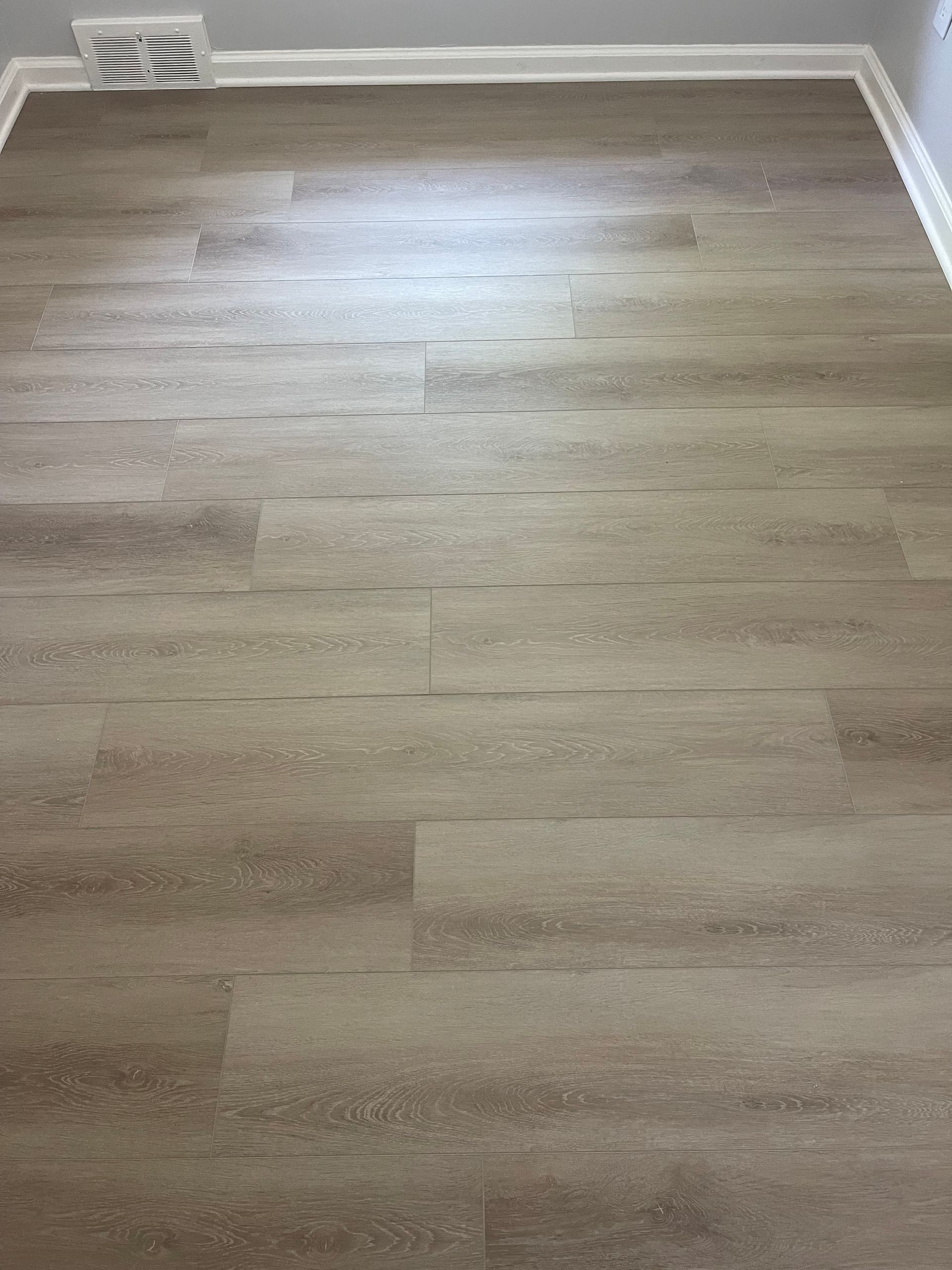 Wooden-look flooring with light brown planks. Air vent in the upper left corner, white trim around the room.