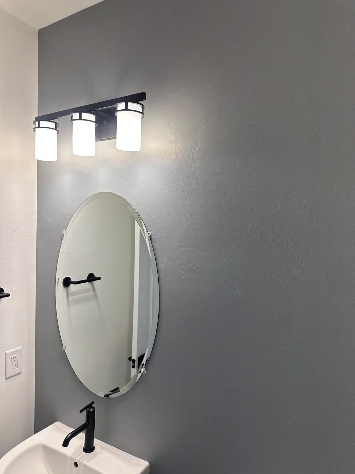 Bathroom with oval mirror, black faucet, and a three-light fixture. The walls are gray and white.