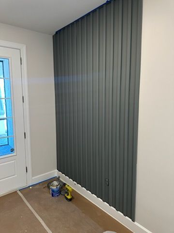 A partially painted accent wall with vertical, ribbed texture, painted gray, next to a white door. Paint cans and tools sit on the floor.