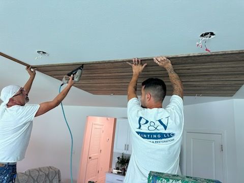 Two men installing wood paneling on a ceiling. One uses a nail gun, the other supports the panel. The room has white walls.