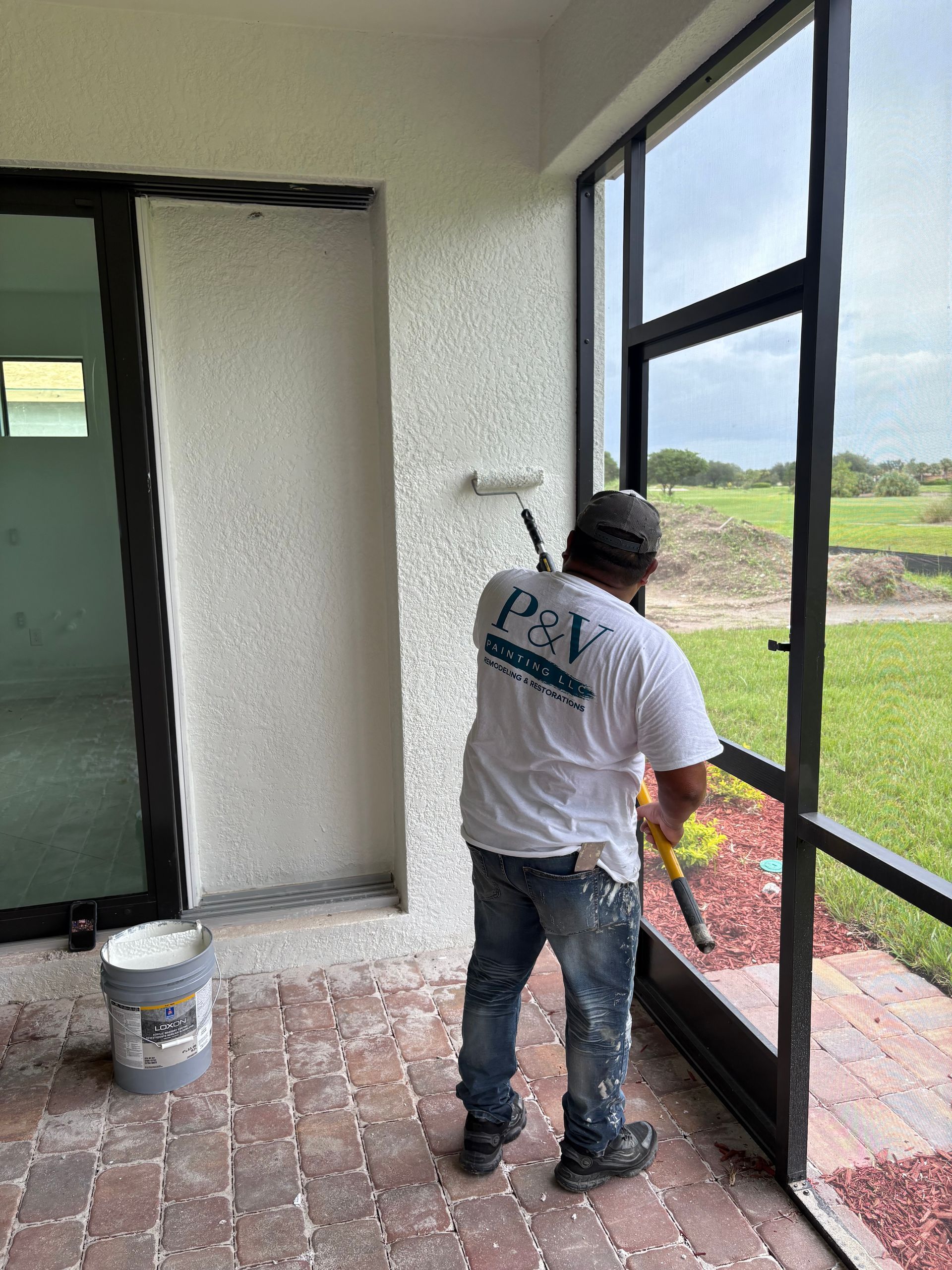 A man in a white shirt and jeans cleans the window frame of a screened-in patio. He stands on a brick patio, working near a grassy field.