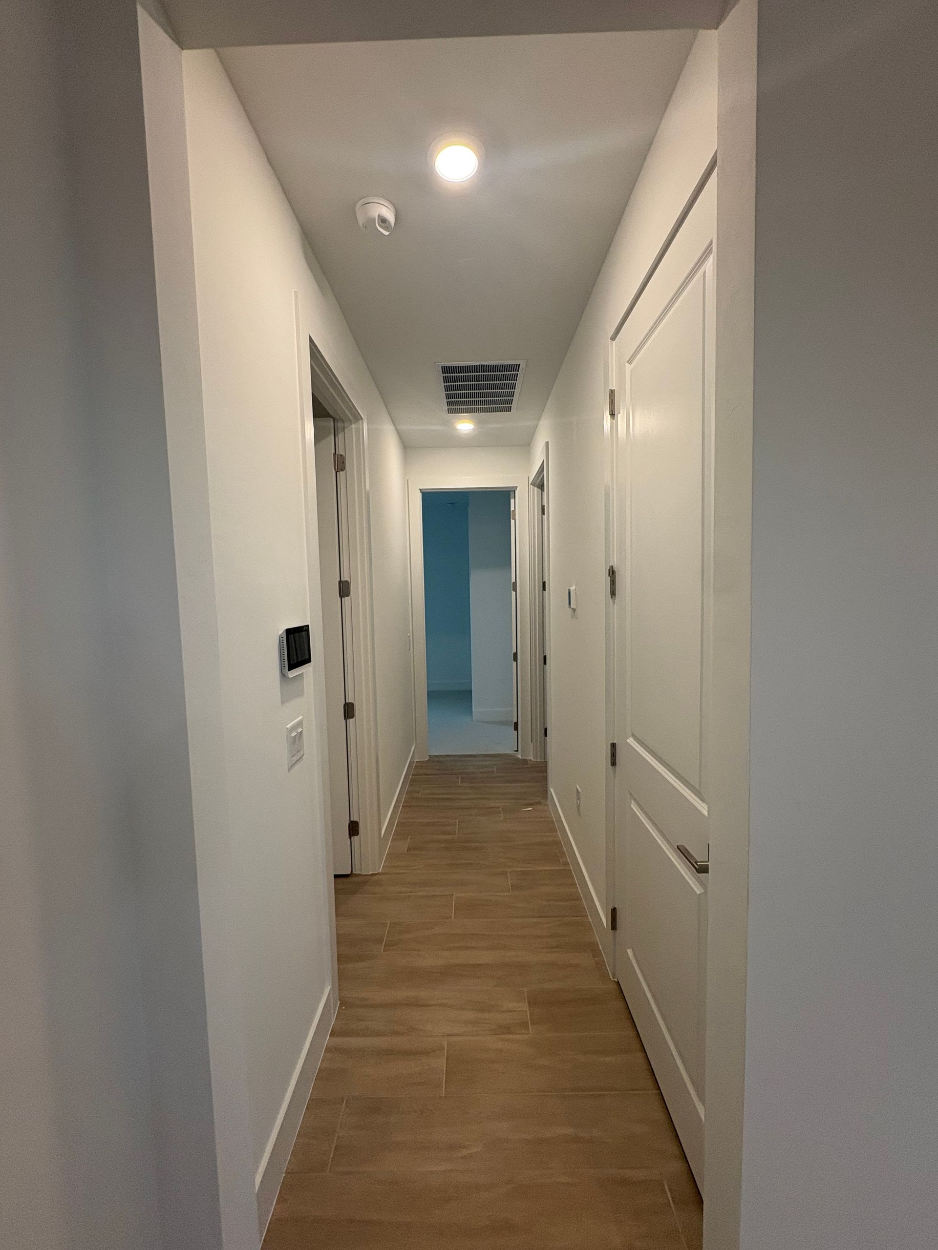 Narrow, white hallway with light brown flooring, several white doors, and a view to a light blue doorway at the end.