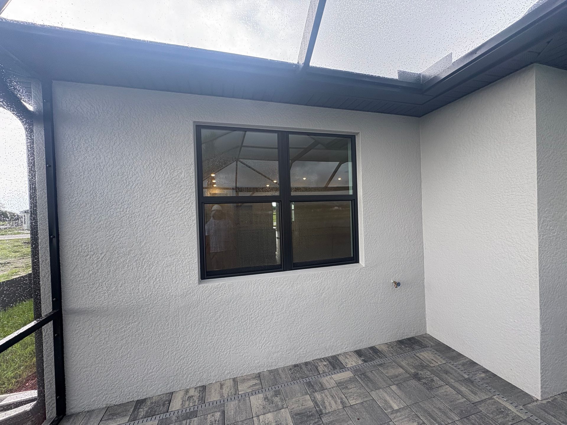 A covered patio with a window. The walls are textured white, with a dark-framed window. The floor is tiled and the roof is clear.