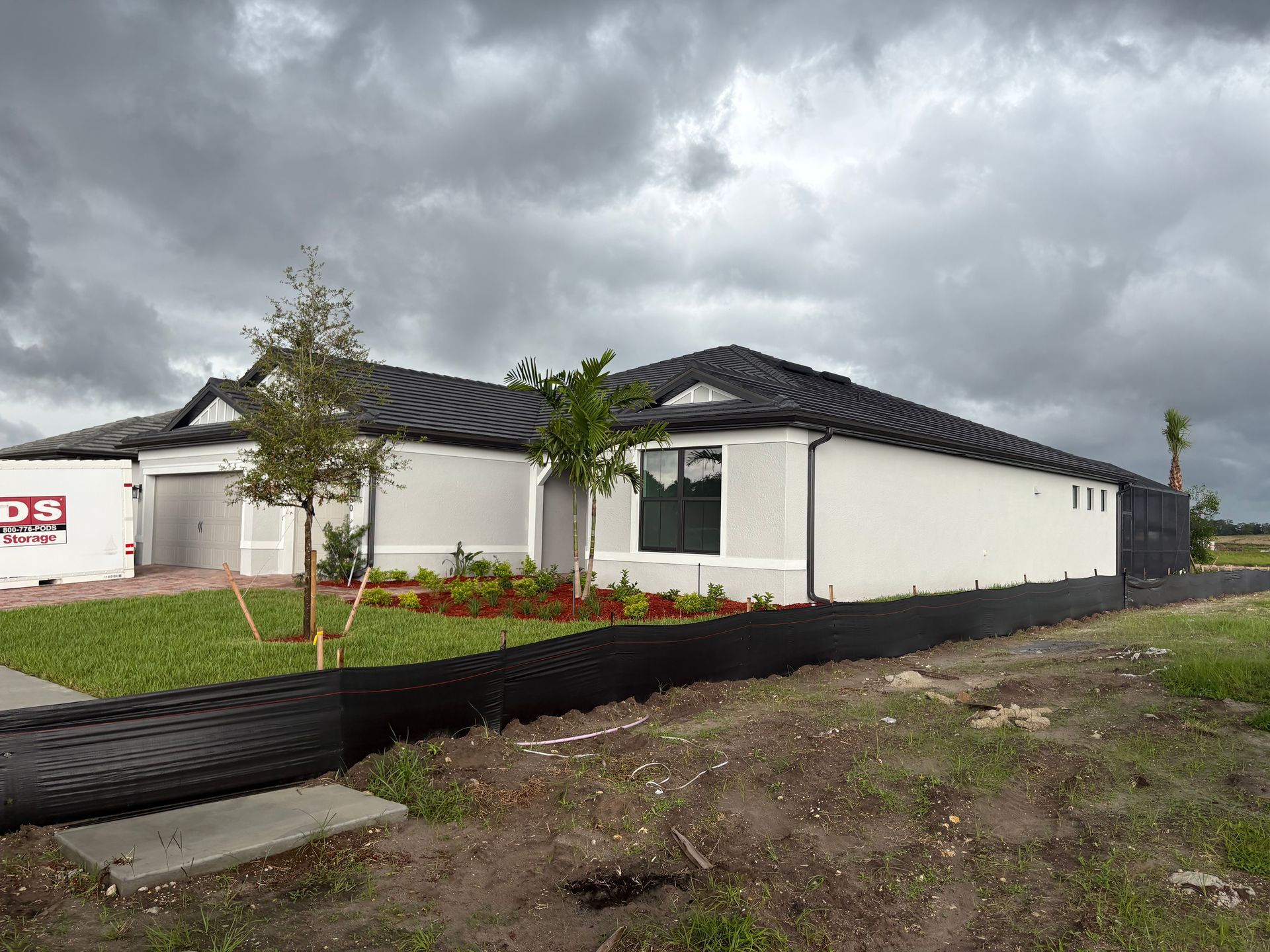 New house under a cloudy sky; light grey exterior, black roof, black silt fence, green lawn with small trees and red flowers.