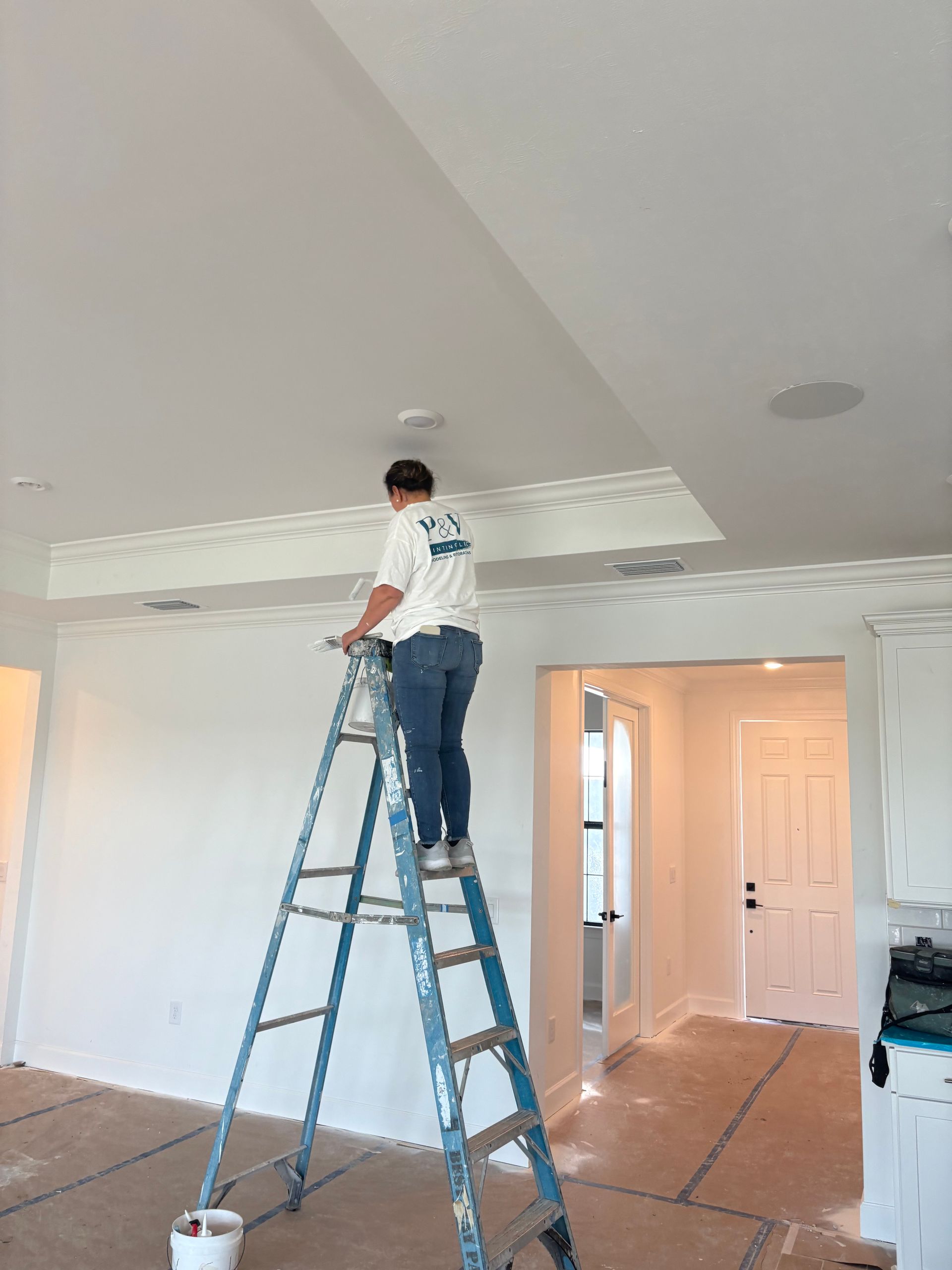 Person on a ladder painting a white ceiling in a newly constructed house.