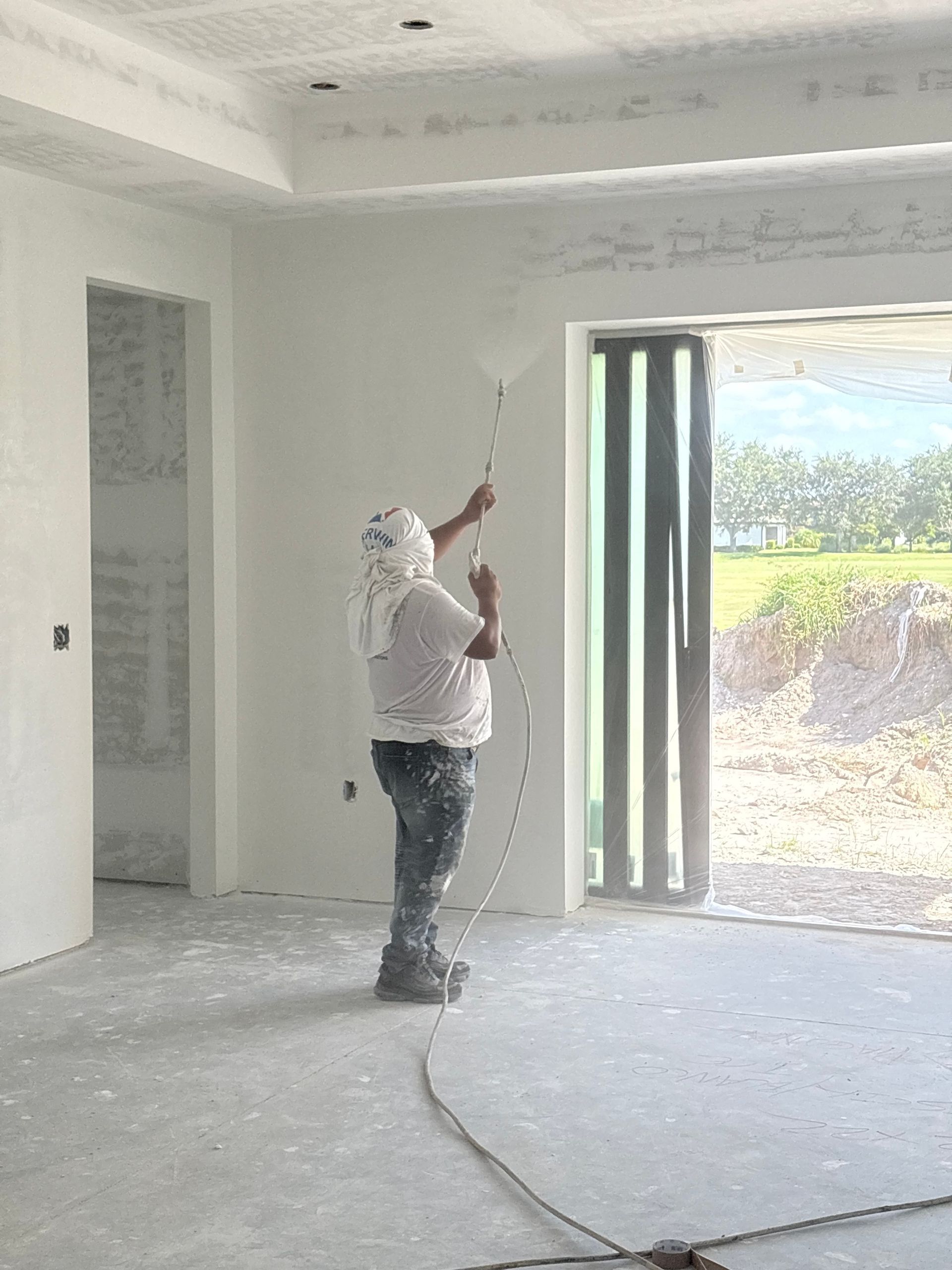 Person spraying a white wall with a paint sprayer inside a room with a sliding glass door, wearing a white shirt and head covering.