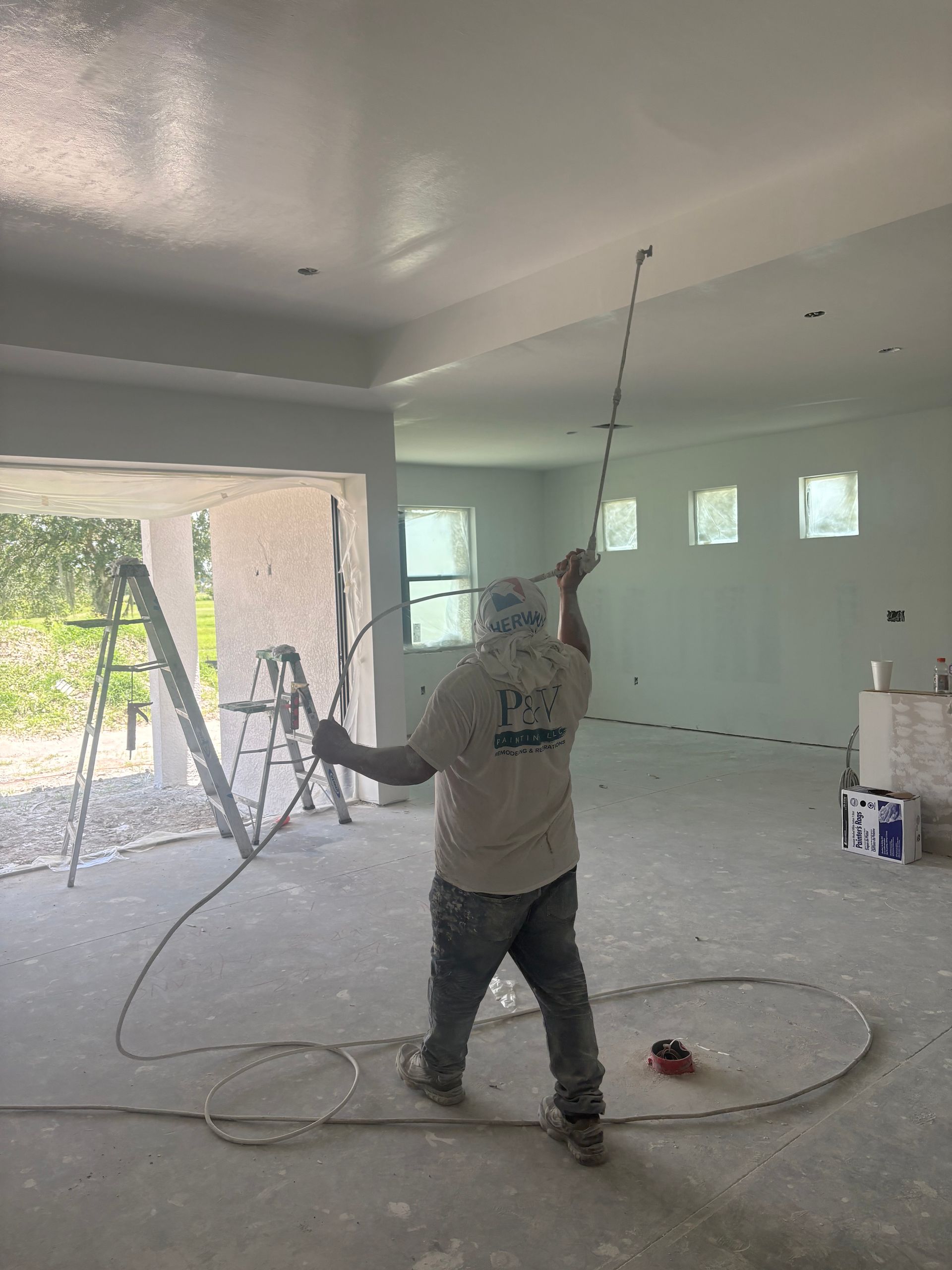A person sprays paint on a ceiling indoors. They wear work clothes and hold a spray gun. The room is mostly white.