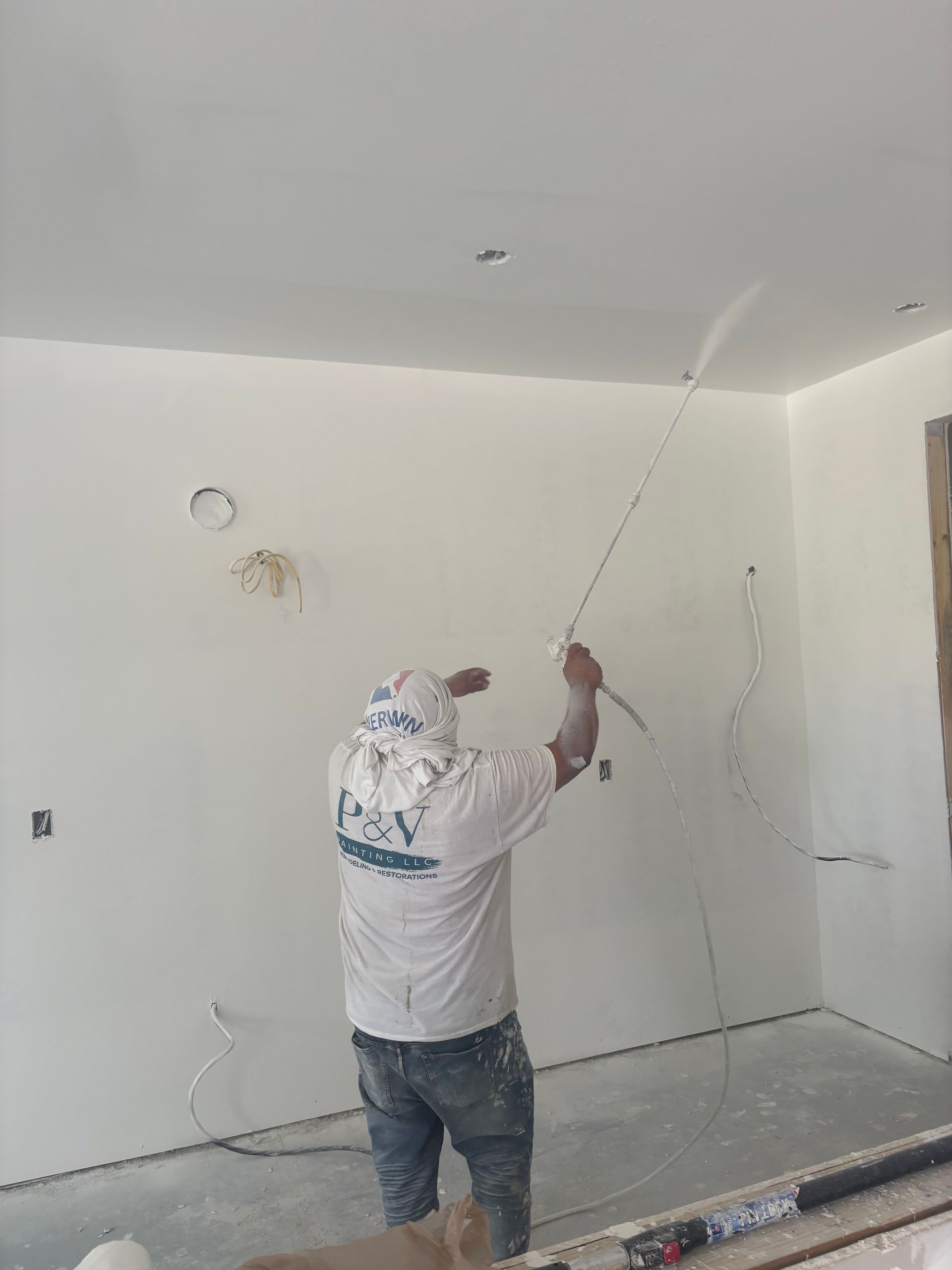 A person is spraying a white substance on a white wall and ceiling in a room with unfinished electrical wiring.