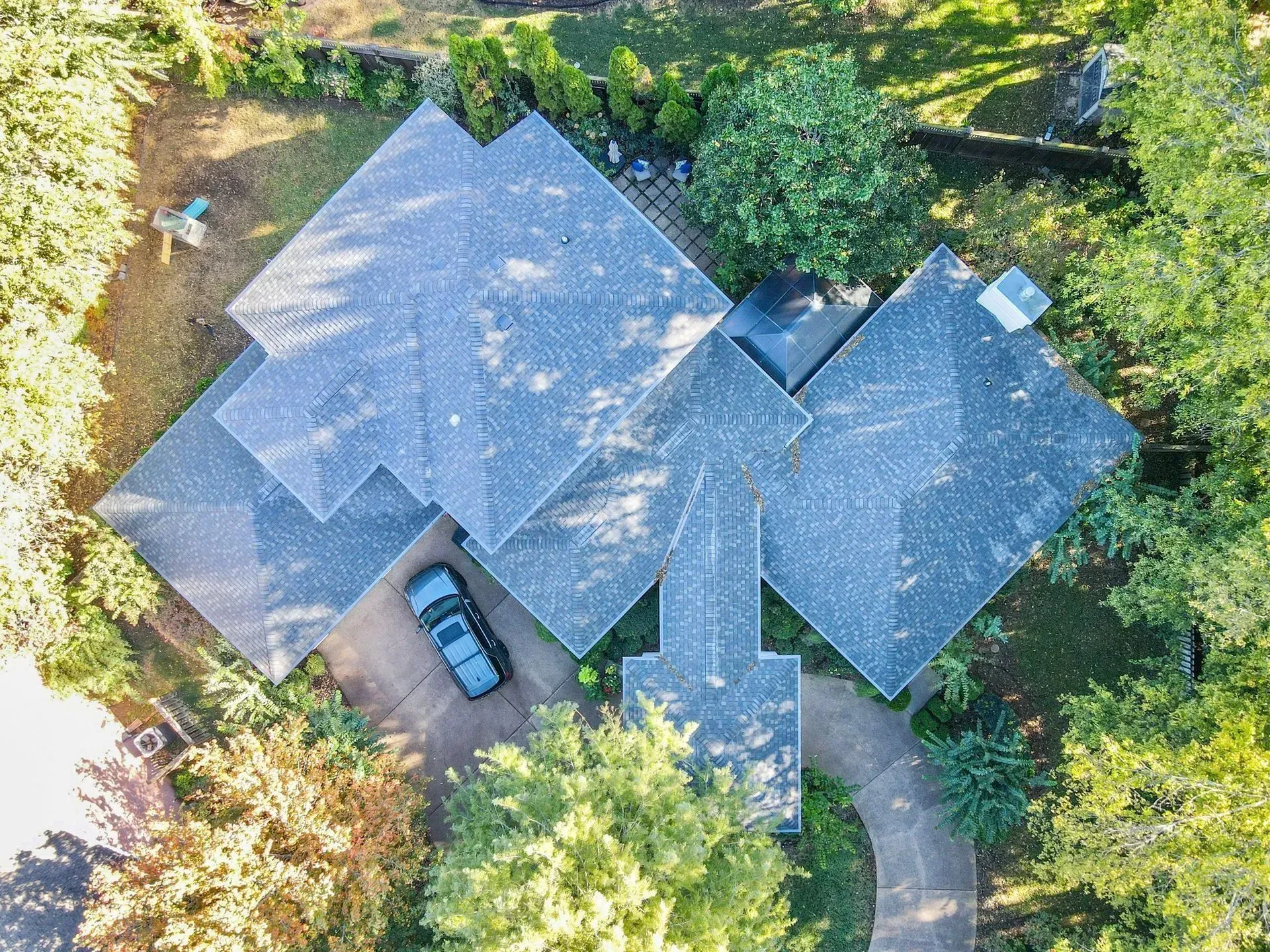 Aerial view of a gray-roofed house with a curved driveway and parked car, surrounded by trees and greenery.