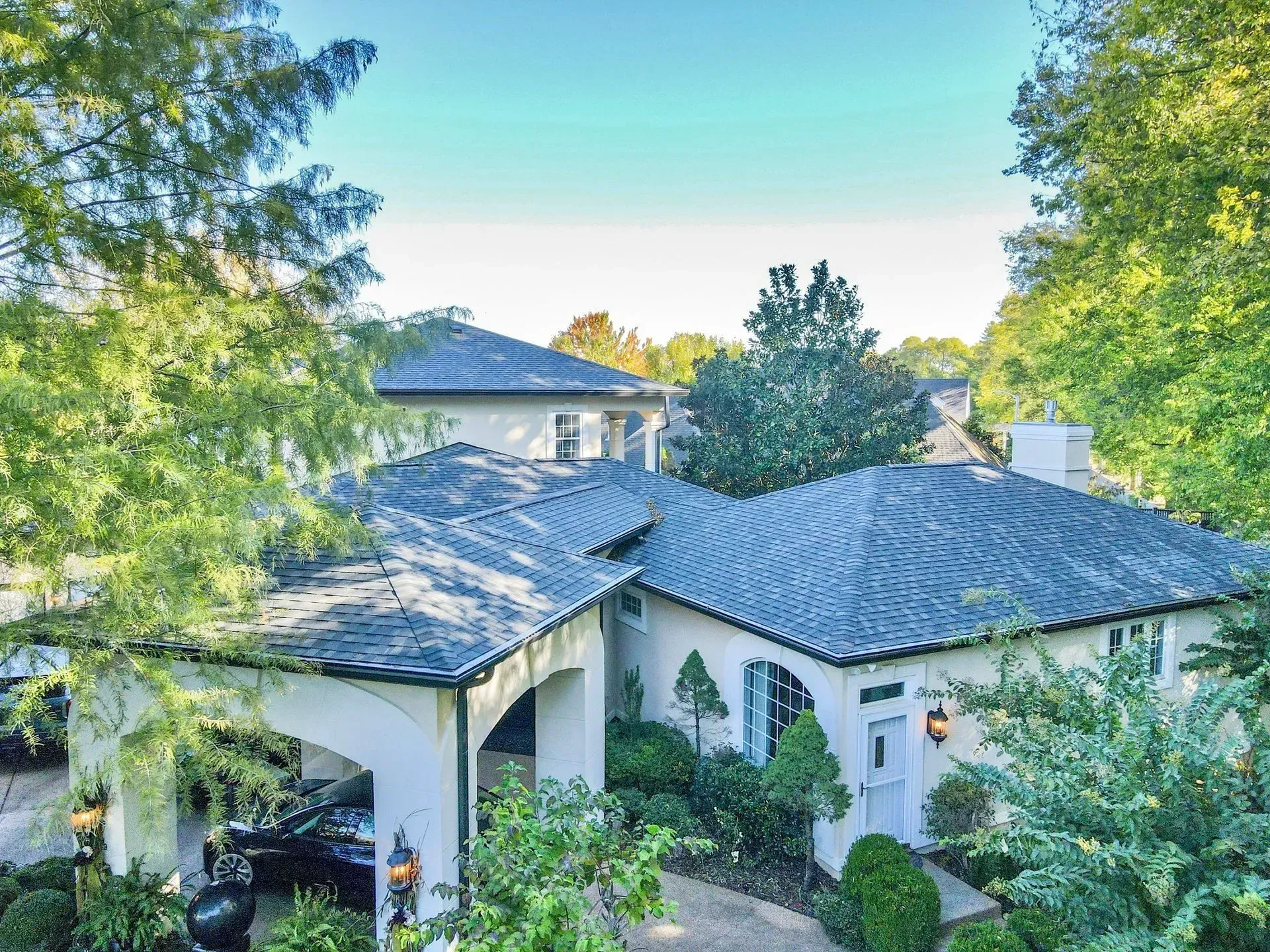 White stucco house with a gray shingle roof surrounded by lush green trees and shrubs.