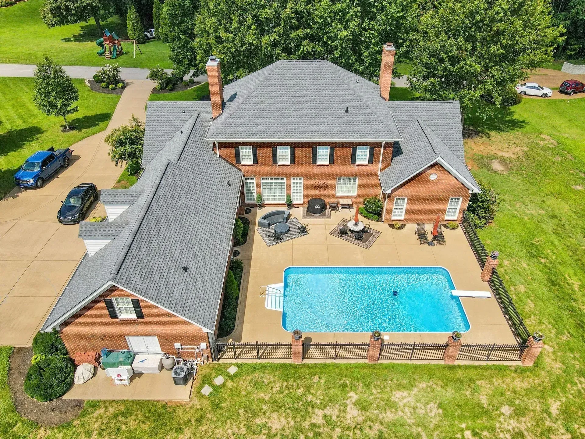 Aerial view of a brick house with a pool, patio, and fenced backyard surrounded by trees.