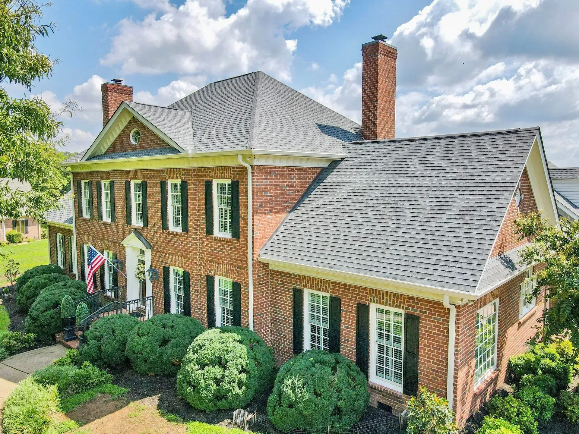Brick house with gray shingled roof, white-trim windows, and manicured green shrubs under a cloudy sky.