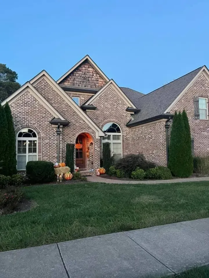Brick suburban house with arched windows, lit front entry, manicured lawn, and driveway at dusk.