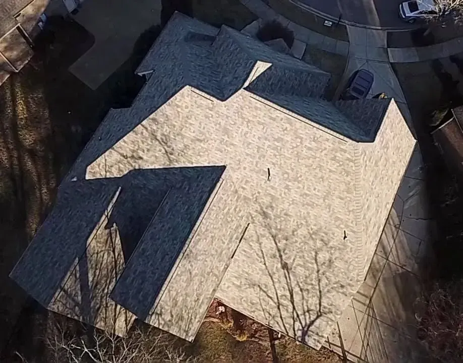 Aerial view of a house with a light roof and dark dormers, surrounded by trees and a driveway.