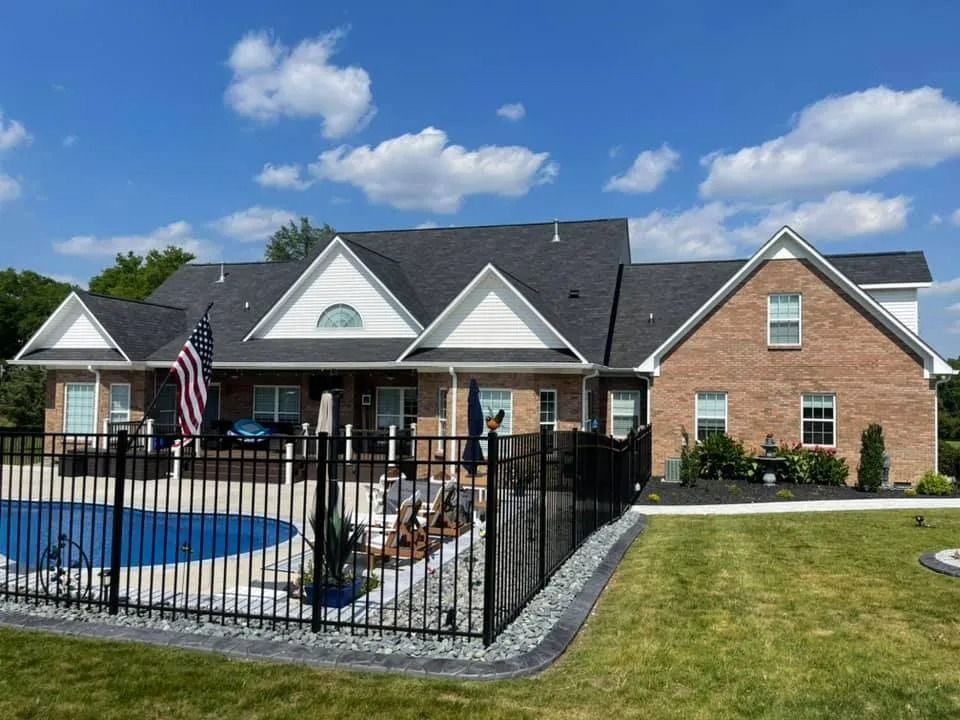 Backyard pool and patio beside a brick house with a black fence under a blue sky