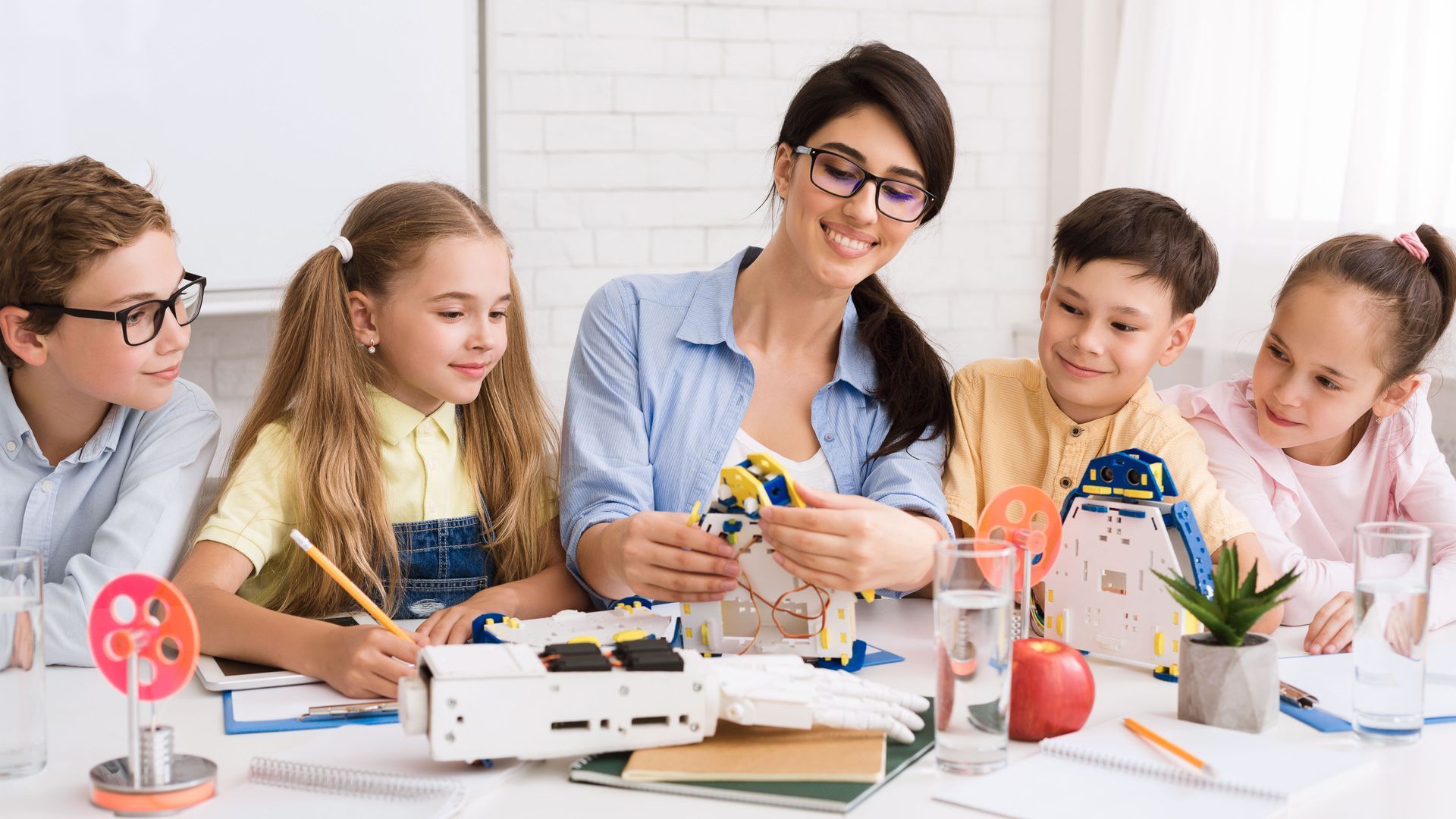 A teacher smiles while demonstrating robotics to four children at a table with robot parts and learning materials.