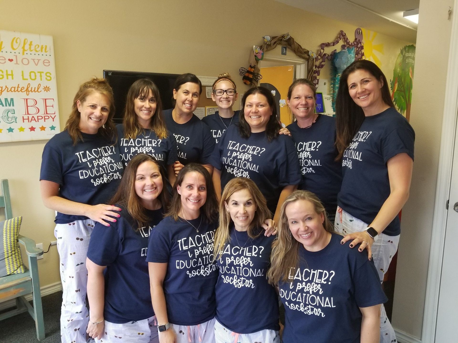 Group of teachers wearing matching navy shirts and pajamas, posing indoors with a wall decoration behind them.