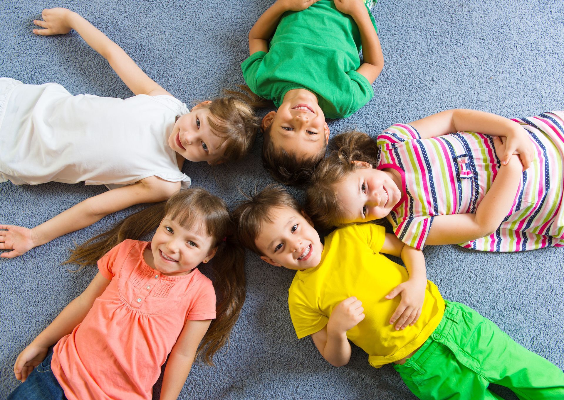 Children in a circle, smiling at the camera. They wear colorful clothes and lie on a blue carpet.