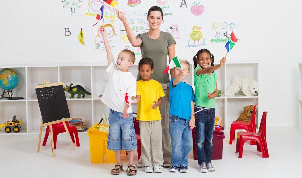 A teacher and four diverse children wave flags in a brightly decorated classroom. Smiling, they stand against a wall with drawings and letters.