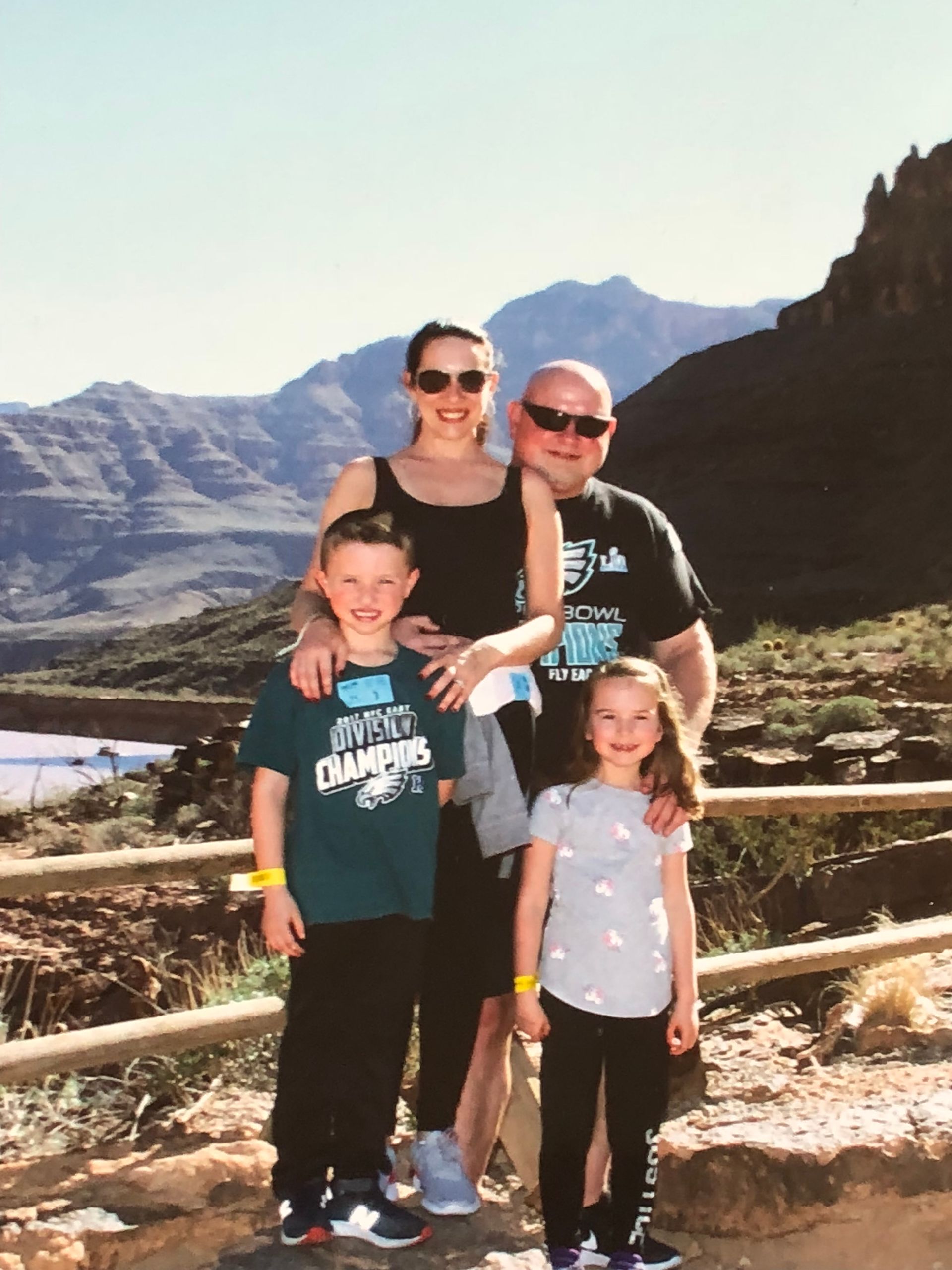 A family of four stands outdoors by a wooden railing with a scenic mountain range in the background.