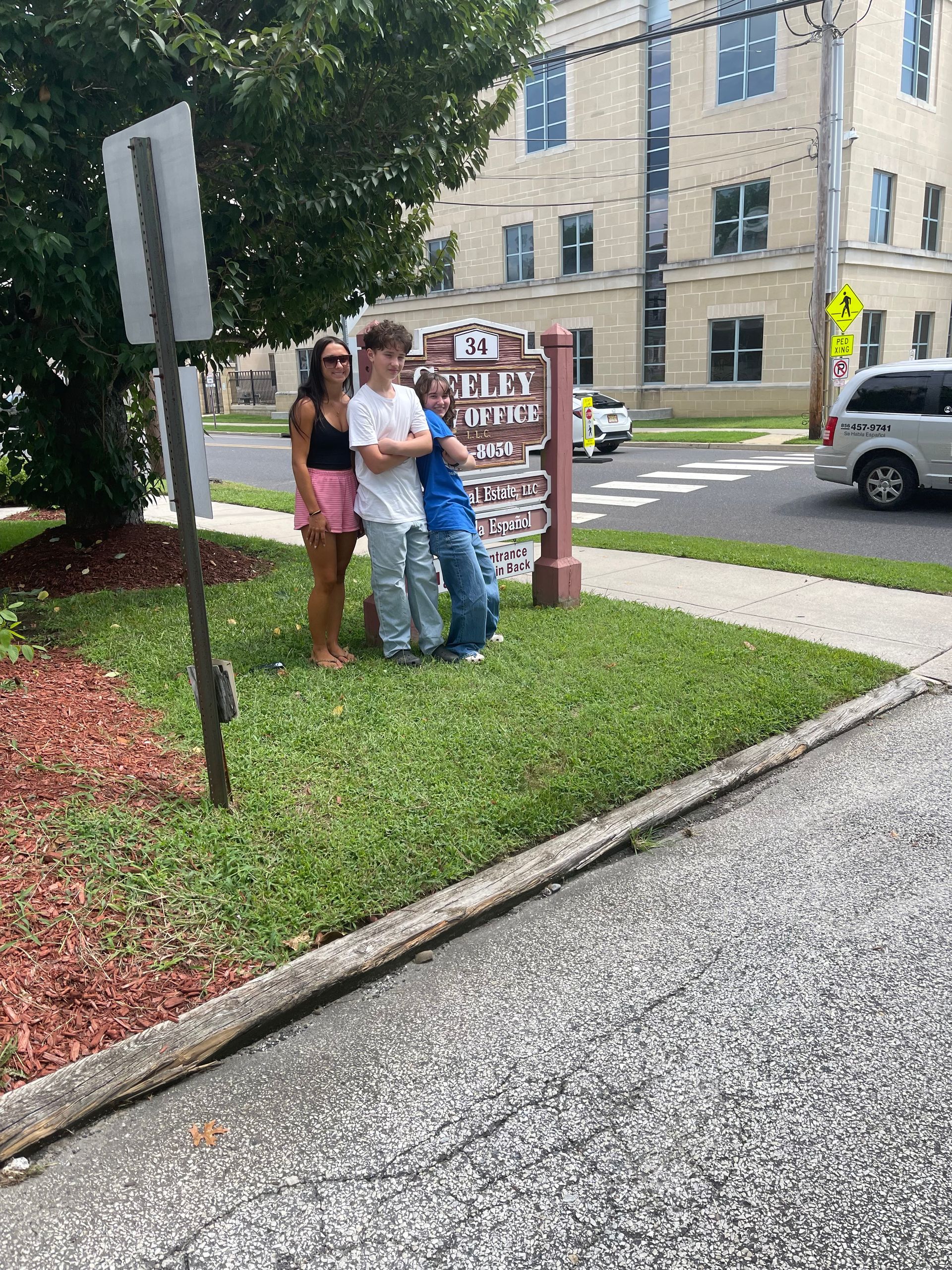 Three people stand together, leaning against a sign for an Elks Lodge on a grassy corner by a street.