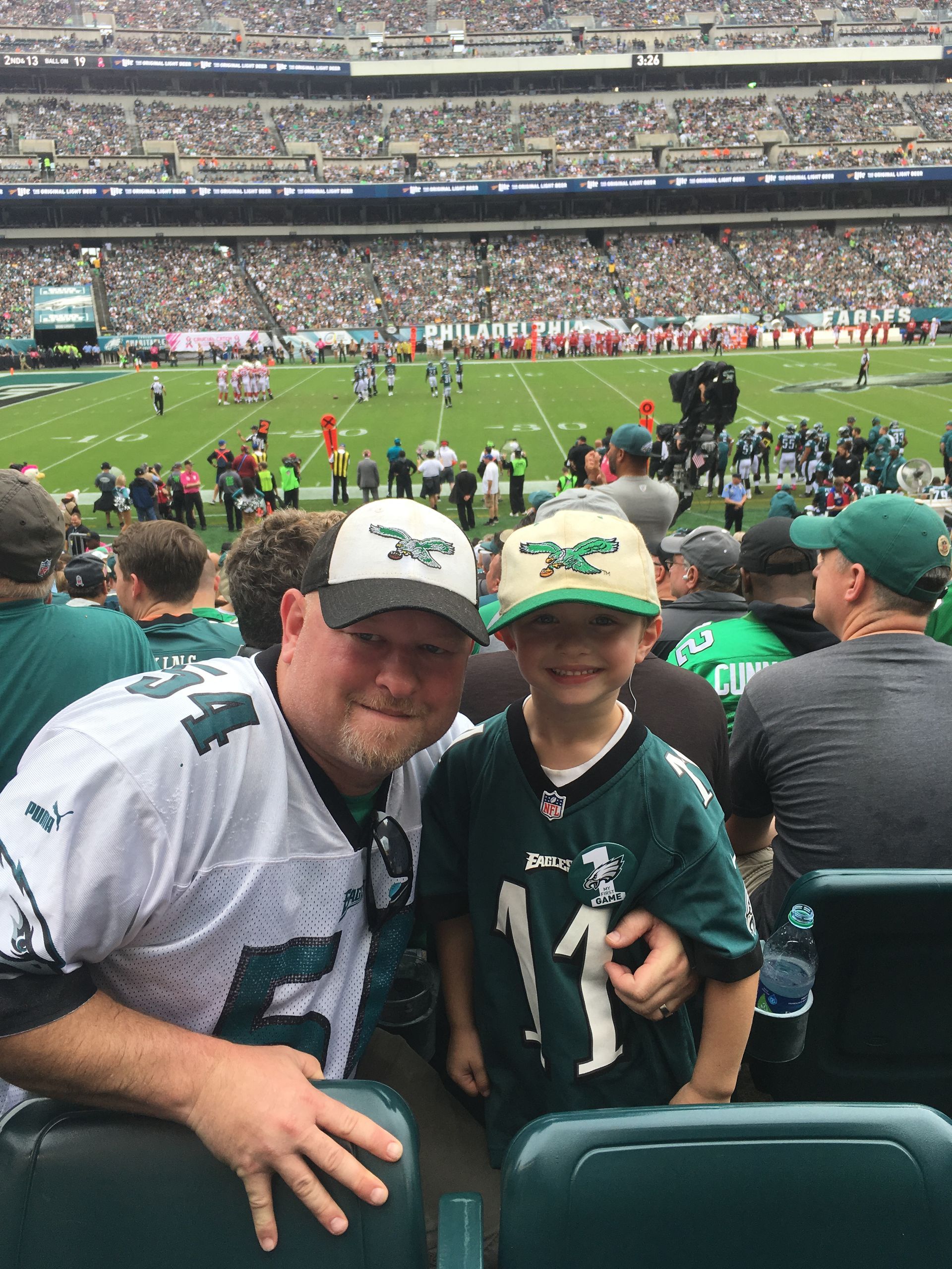 A fan and child wearing Philadelphia Eagles gear smile for a selfie at a football stadium during a game.