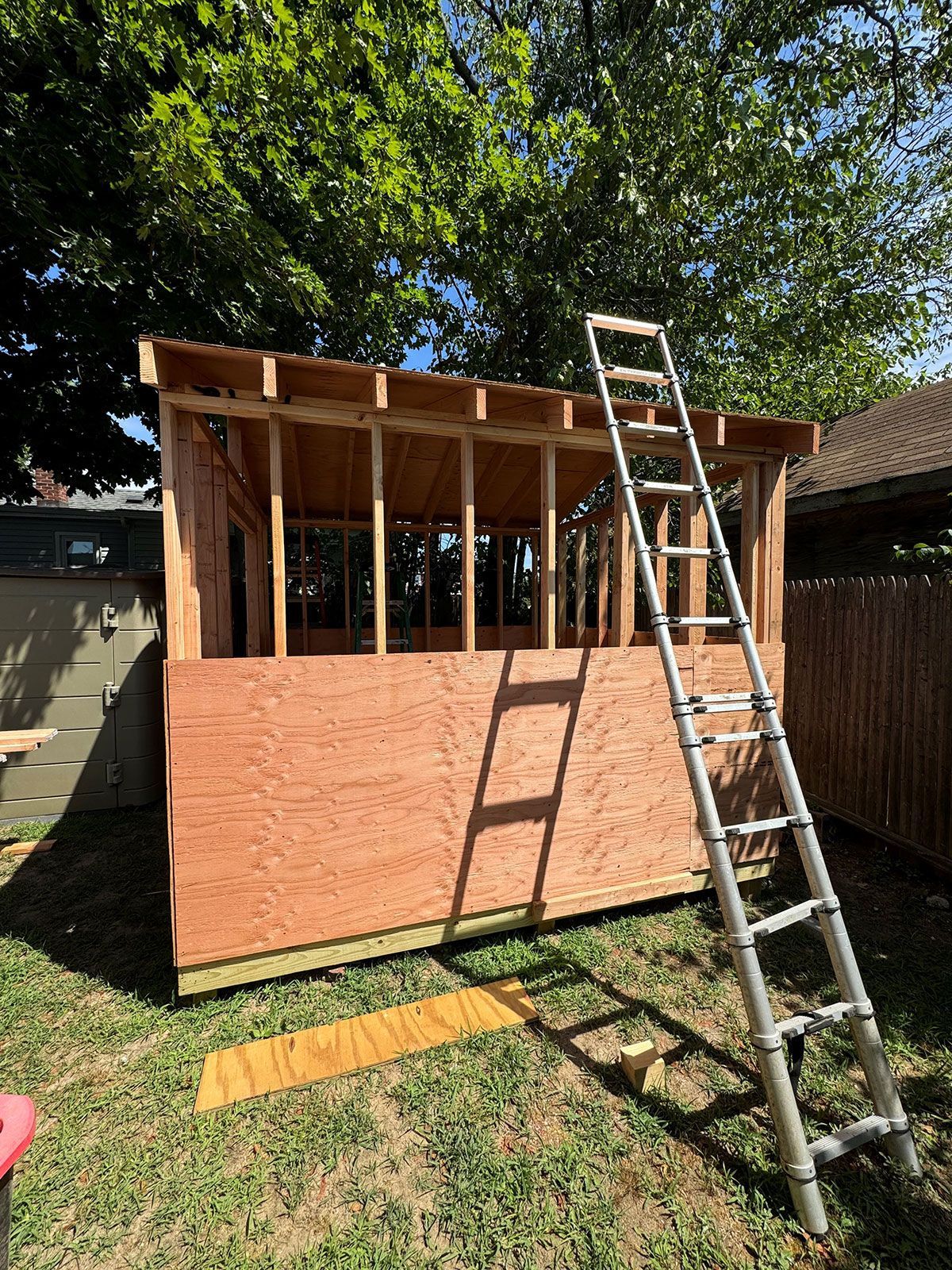 A wooden shed is being built in a backyard with a ladder attached to it.