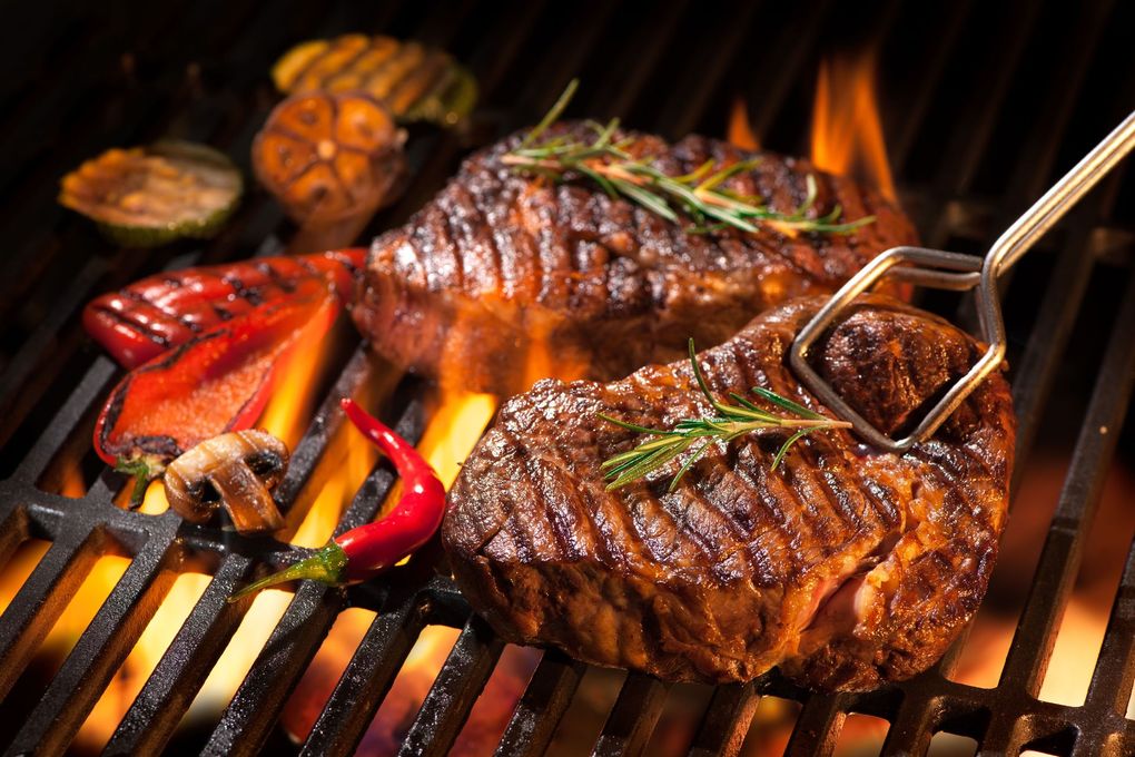 Two grilled steaks with herbs on a barbecue grill, surrounded by flames and vegetables