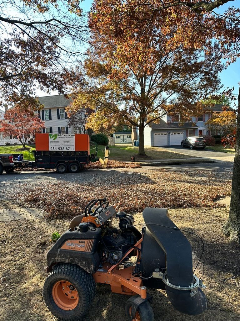 Lawnmower collecting fallen autumn leaves; truck parked nearby. Residential neighborhood with colorful trees and houses.