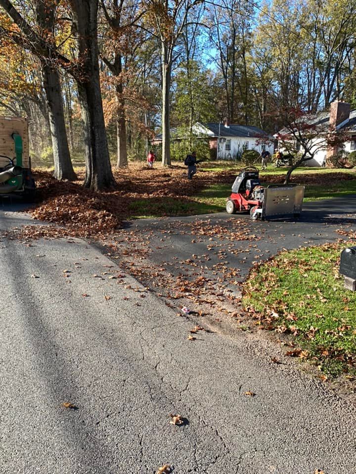 Workers clearing fallen autumn leaves from a driveway with a leaf vacuum, in front of houses and trees.