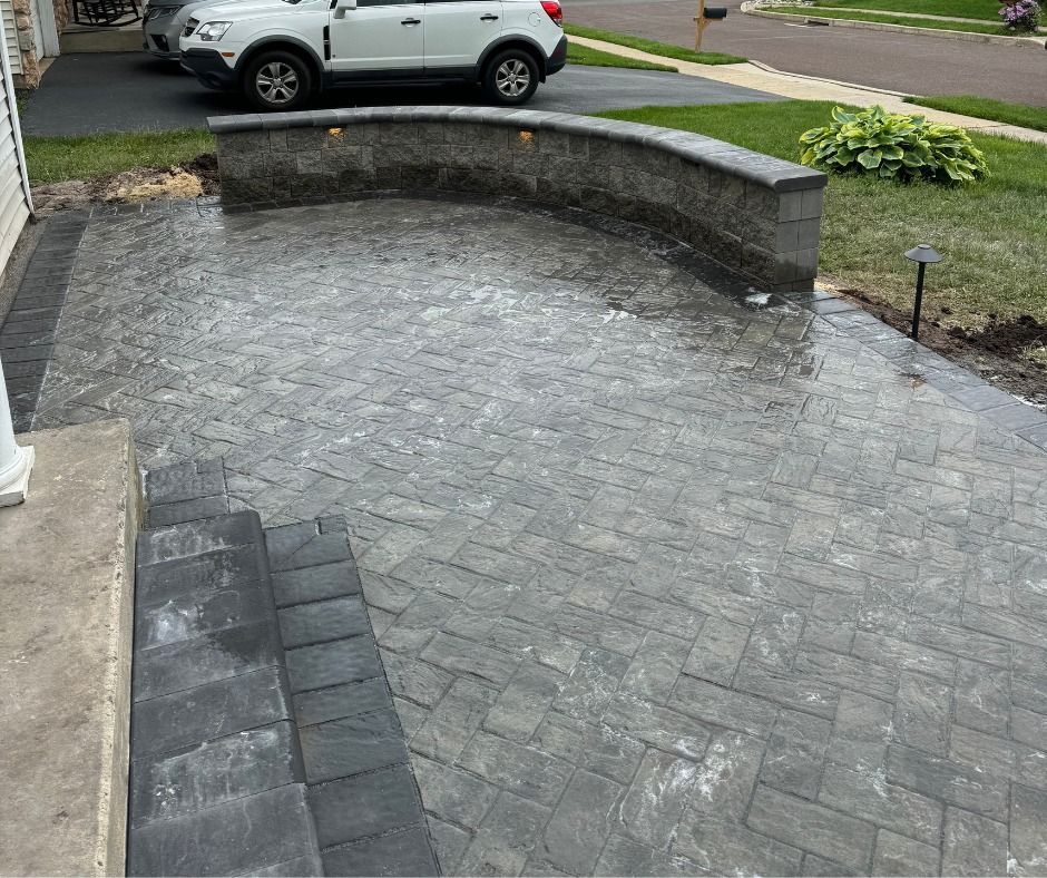Gray brick paved patio with retaining wall, steps leading up. Car parked in driveway.