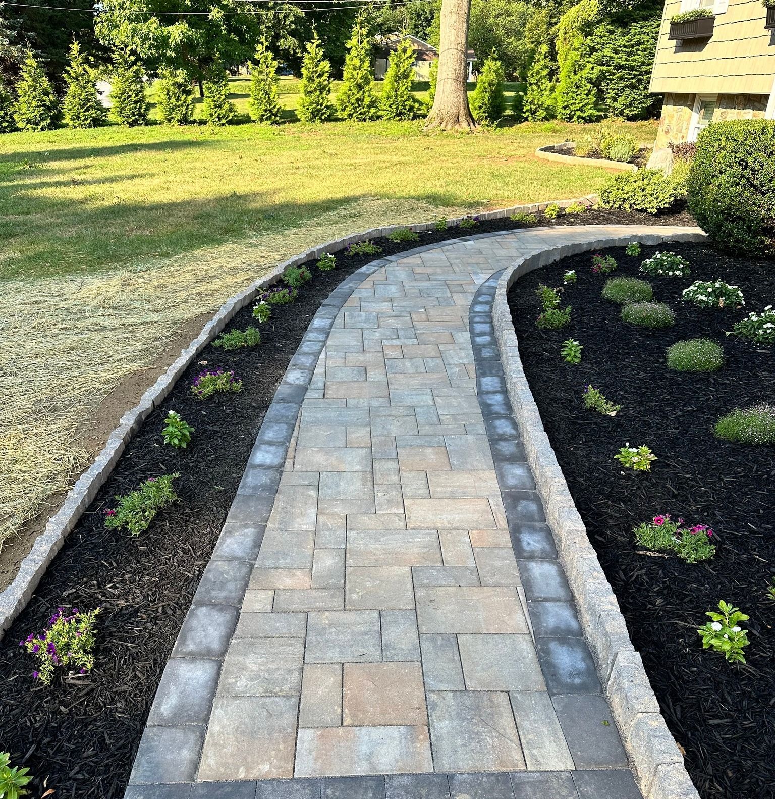 Stone path curves through a landscaped garden with dark mulch and small plants; lawn in background.