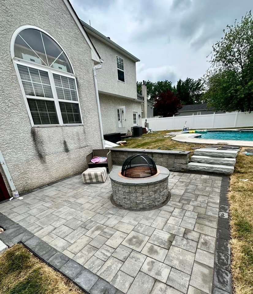Stone patio with a fire pit, next to a house with a pool in the background.