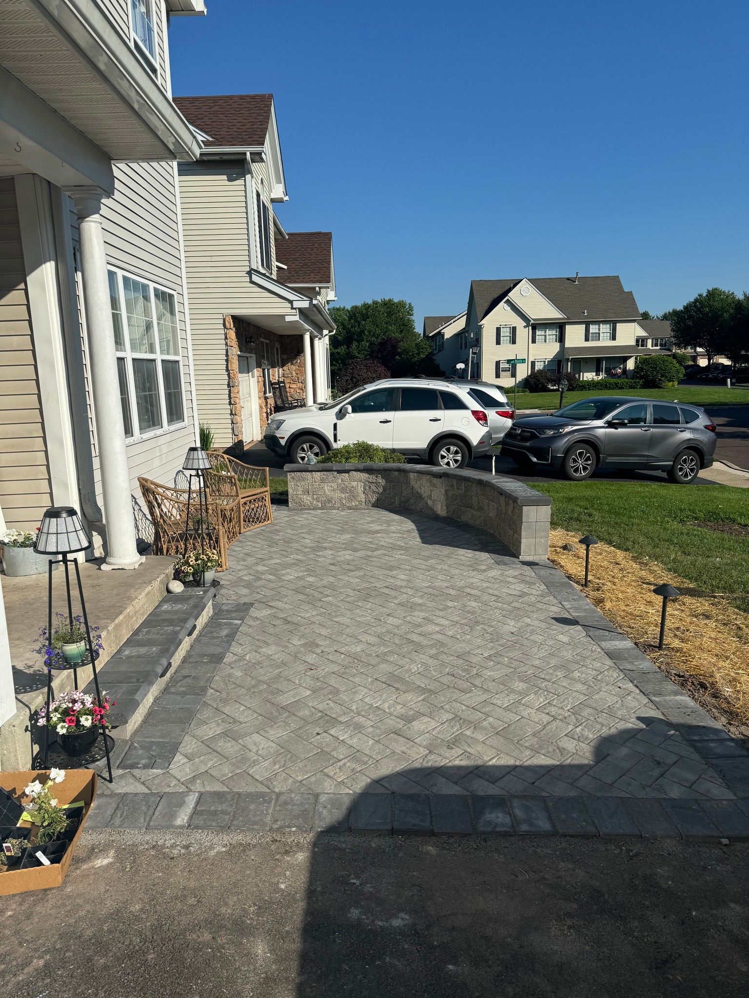 Stone walkway leading to a house with cars parked nearby, under a blue sky.