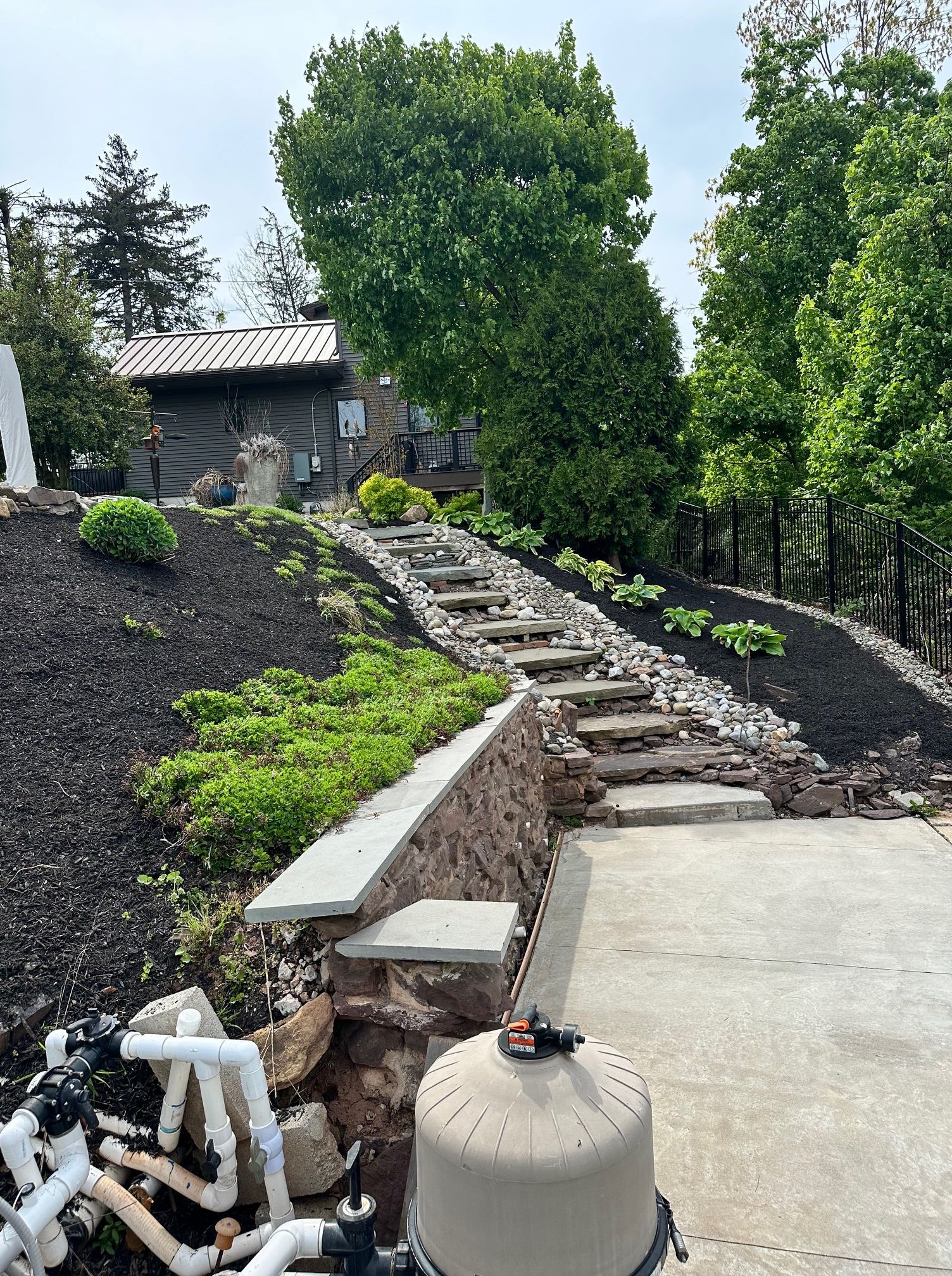 Stone steps lead up a landscaped hill with dark mulch and greenery; pool equipment in foreground.